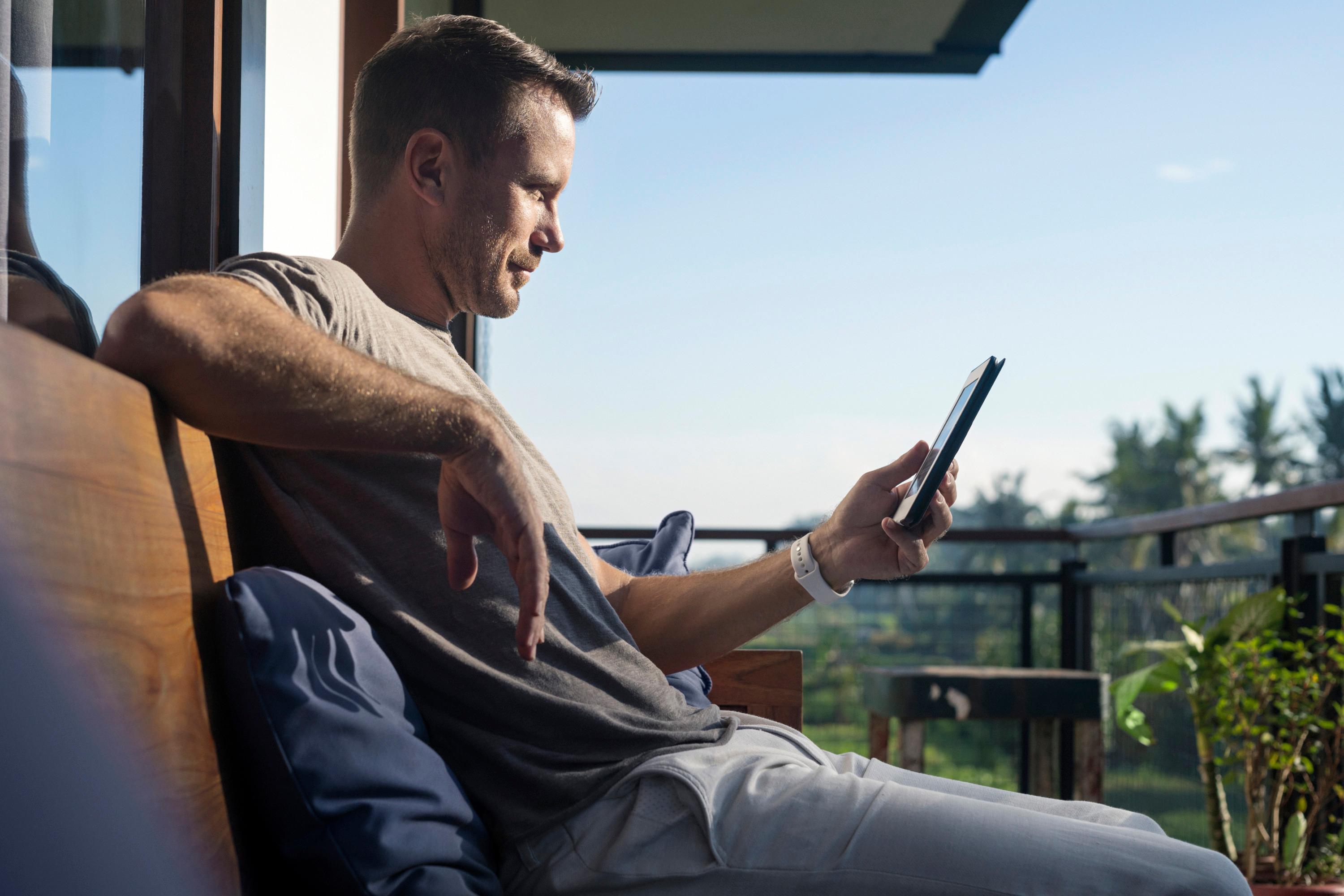 HE - Image of a man sitting on a balcony looking at his phone with a green field outside. He is smiling at his phone, comfortable knowing that he can remote monitor his home's power status.