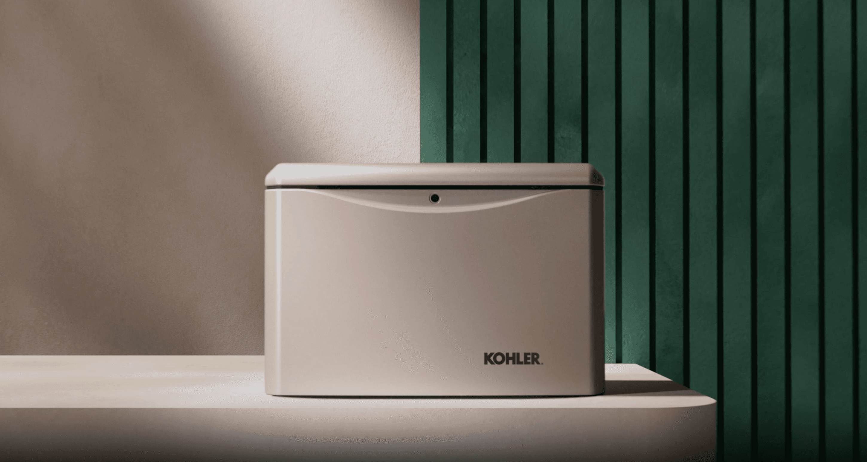 Artful image of grey Kohler home generator on a rectangle platform with rounded corners. Behind the generator is a wall of narrow wood panel painted green. Dramatic lighting casts shadows across the platform and generator